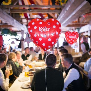 Guests at long tables in an Oktoberfest tent under a red heart balloon