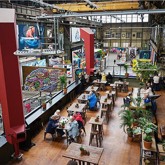 People at long tables in a converted Amsterdam warehouse hung with murals